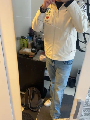 Vit vindjacka Brasil med dragkedja - Snygg vit vindjacka från Nike med Brasil-tryck och broderade detaljer på bröstet. Jackan har hel dragkedja framtill, hög krage och lång ärm. Perfekt för dig som gillar sportig stil och vill sticka ut med coola detaljer. Pris kan diskuteras