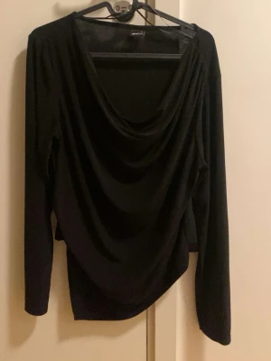 Svart draperad långärmad topp - Svart långärmad topp från Gina Tricot med snygg draperad urringning. Toppens material är mjukt och stretchigt, vilket ger en bekväm passform. Perfekt för dig som gillar stilrena och trendiga plagg.