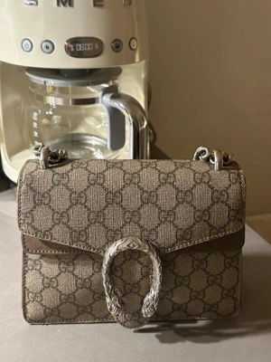 Gucci Dionysus axelväska monogram - Snygg axelväska från Gucci i klassiskt GG-monogrammönster. Väskan är brun med silverfärgad kedja och ikoniskt spänne i form av ett tigerhuvud. Tillverkad i canvas med läderdetaljer. Perfekt för dig som vill ha en trendig och lyxig accessoar.