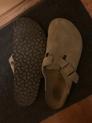 Beige mockatofflor från Birkenstock - Snygga beige tofflor från Birkenstock i mocka med klassisk täckt tå och justerbart spänne i metall. Mjuk innersula och räfflad gummisula för bra grepp. Perfekta för chill dagar hemma eller när du vill ha det bekvämt. Knappt använda eftersom de är lite stora för mig.