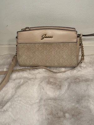 Beige axelväska från Guess - Snygg beige axelväska från Guess med guldfärgad logga framtill. Väskan har ett mönstrat tyg med diskreta G-detaljer och ljusbeiga läderdetaljer upptill. Justerbar axelrem i läder och kedja. Perfekt för att bära det viktigaste med stil.
