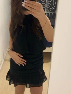 Svart kort klänning med volang - Snygg svart kort klänning med volang nertill och ärmlös design. Klänningen har en lätt rynkad effekt vid höften och är tillverkad i ett tunt, flowy material som ger en elegant känsla. Passar 34/36