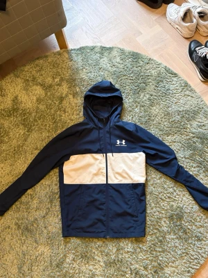 Under armour windbreaker - Snygg hoodie från Under Armour i marinblått med vit panel över bröstet och dragkedja framtill. Jackan har huva och två fickor framtill. Perfekt för en sportig look och tillverkad i ett lätt och smidigt material.