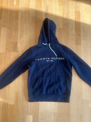 Marinblå hoodie Tommy Hilfiger - Snygg marinblå hoodie från Tommy Hilfiger med broderad logga och text på bröstet. Klassisk känguruficka framtill och vit dragsko i huvan. Perfekt för en avslappnad och stilren look.