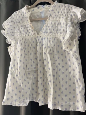 Vit blus med volangärmar och blått mönster - Söt vit blus med små blå blommönster, volangärmar och v-ringning. Blusen har rynkade detaljer upptill och är tillverkad i ett lätt, luftigt tyg som känns perfekt för varma dagar. Passar dig som gillar romantisk och bohemisk stil.