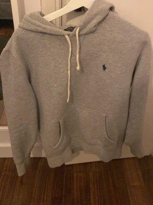 Grå hoodie från Polo Ralph Lauren - Snygg grå hoodie från Polo Ralph Lauren med klassisk huva och vita dragsnören. Har en känguruficka framtill och den ikoniska lilla blå loggan broderad på bröstet. Tillverkad i mjukt sweatshirtmaterial som är perfekt för chill dagar.