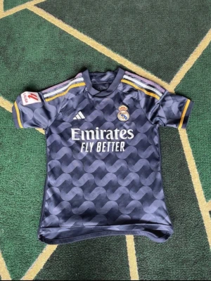 Real Madrid matchtröja Adidas - Snygg mörkblå Real Madrid matchtröja från Adidas med gul och vit detalj på axlarna. Tryck med Emirates Fly Better på bröstet och La Liga-märke på ärmen. Tillverkad i lätt och ventilerande material, perfekt för fotbollsträning eller match.