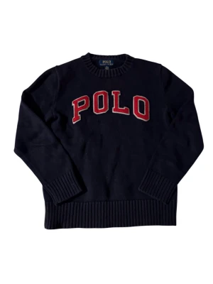 Mörkblå stickad tröja Polo Ralph Lauren - Mörkblå stickad tröja från Polo Ralph Lauren med stor röd och vit POLO-text framtill. Tröjan har rund halsringning, ribbstickade muddar och nederkant. Perfekt för dig som gillar klassisk college-stil.