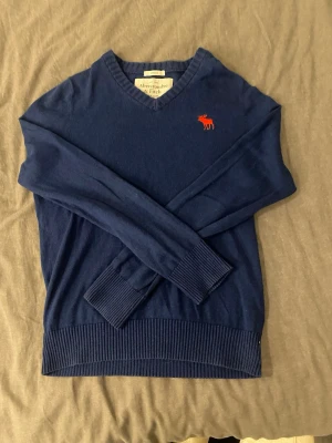 Mörkblå stickad tröja Abercrombie - Mörkblå stickad tröja från Abercrombie & Fitch med V-ringning och röd broderad älglogga på bröstet. Tröjan har ribbstickade muddar vid ärmslut och nederkant. Perfekt för dig som gillar klassisk och stilren design.
