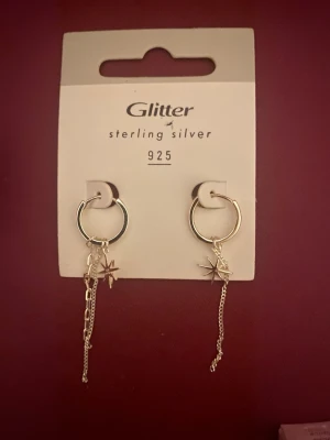 Silvriga örhängen med stjärnhänge Glitter - Snygga örhängen i äkta sterling silver 925 från Glitter. Runda hoops med hängande kedjor och stjärnformade hängen som ger en trendig och cool look. Perfekta för att lyfta din stil med lite extra edge och glans. Oanvända💕💕💕💕