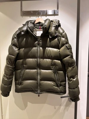 Olivgrön dunjacka från Moncler - Snygg olivgrön dunjacka från Moncler med klassisk puffer-design och huva. Jackan har två dragkedjeförsedda fickor framtill och ikonisk Moncler-logga på ärmen. Tillverkad i glansigt material som ger en exklusiv look. Perfekt för kalla vinterdagar. Perfekt till dig som vill ha en jacka till hösten utan kofta under. Skriv gärna vid frågor😀