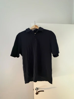 Svart pikétröja från A Day's March - Klassisk svart pikétröja från A Day's March med krage och knappar framtill. Tröjan har korta ärmar och är tillverkad i ett mjukt bomullsmaterial. Perfekt till jeans eller shorts för en stilren look.