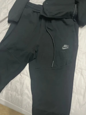 Svart Nike Tech Fleece hoodie - Säljer en svart Nike Tech Fleece hoodie med dragkedja och huva. Hoodien har en snygg detalj med en svart rand över bröstet och en ficka med dragkedja på ärmen. Tillverkad i mjukt och lätt material som är perfekt för chill eller träning.