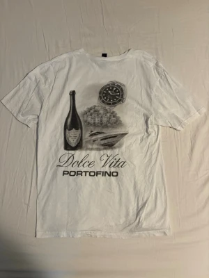 Vit Dolce Vita Portofino t-shirt - Vit t-shirt med tryck på framsidan som visar en flaska, klocka, båt och Portofino-motiv samt texten 'Dolce Vita Portofino'. Klassisk passform med rund hals och korta ärmar. Perfekt för dig som gillar stilrena och unika tryck.