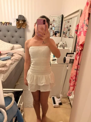 Vit axelbandslös kortklänning - Söt vit axelbandslös kortklänning med smockad överdel och volang nedtill. Klänningen har en figurnära passform upptill och en lösare kjol. Perfekt för varma dagar och enkel att styla med accessoarer.