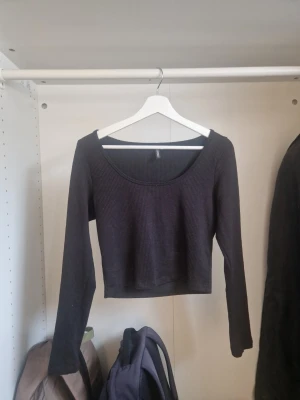 Svart ribbad crop top med lång ärm - Trendig svart ribbad crop top med långa ärmar och bred, rundad urringning. Toppen är figurnära och har en kortare passform som passar perfekt till höga jeans eller kjol. Materialet är stretchigt och mjukt för en bekväm känsla.