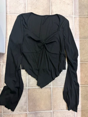 Svart långärmad topp med knut - Trendig svart topp med lång ärm och snygg knutdetalj framtill. Toppen har en asymmetrisk nederkant och croppad passform som ger en edgy look. Perfekt för dig som vill sticka ut lite extra.