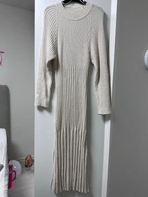 Gräddvit ribbad stickad långklänning - Snygg beige långklänning i ribbstickat material med lång ärm och rund halsringning. Klänningen har en figurnära passform och är perfekt för dig som gillar en stilren och mysig look.
