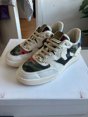 Valentino camo sneakers i läder - Snygga sneakers från Valentino med beige läder och camo-mönstrade paneler i grönt, svart och rött. Skorna har vita sulor, beige skosnören och en detalj i blått på hälen. Märkeslogga på plösen och cool nitar-detalj vid snörningen.