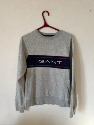 Grå sweatshirt från GANT - Snygg grå sweatshirt från GANT med marinblå panel och röd detalj över bröstet samt stor logga framtill. Tröjan har rund halsringning, långa ärmar och ribbade muddar. Perfekt för en avslappnad och stilren look.