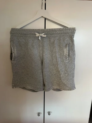 Grå mjukisshorts med snörning - Mysiga grå shorts i mjuk bomullsblandning med elastisk midja och vit snörning framtill. Två fickor på sidorna och en bakficka. Dock är det hål i båda sidofickorna (Se bild)!