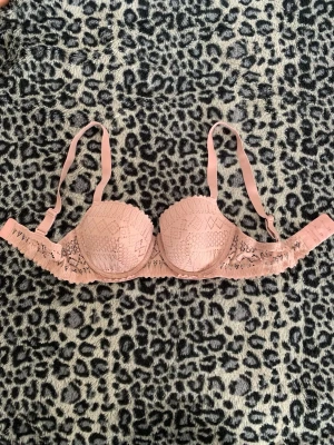 Rosa spets-bh med vadderade kupor - Söt bh i ljusrosa med vadderade kupor och dekorativ spets framtill. Justerbara axelband och hakknäppning bak. Kuporna har ett diskret mönster och sidorna är i genombruten spets för en feminin look. Perfekt för dig som gillar romantiska detaljer.
