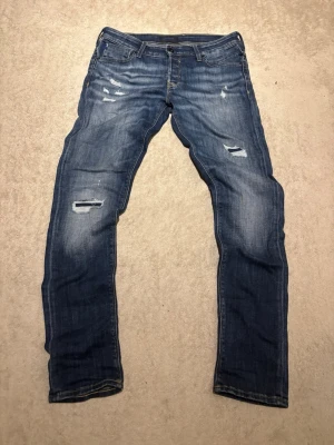Blå slitna jeans med raka ben - Säljer ett par blå jeans med slitningar och hål på låren och knäna. Jeansen har raka ben, klassisk femficksmodell och detaljerade sömmar. Perfekta för en avslappnad och trendig look.