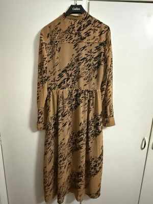 Beige och svart långklänning från Calvin Klein -  En stilren långklänning från Calvin Klein i beige med ett svart, abstrakt mönster som verkligen sticker ut. Klänningen har en smalare passform som framhäver silhuetten, långa ärmar och knäppning framtill. Den är dessutom utrustad med en dragkedja på vänster sida för enkel av- och påklädning, samt praktiska sidofickor. Perfekt för dig som gillar unika plagg med en modern och självsäker stil.