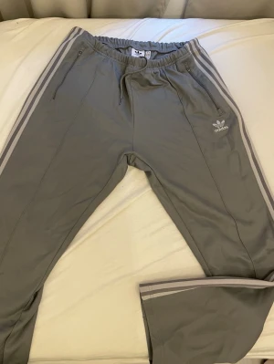 Adidas mjukisbyxor  - Säljer ett par grå mjukisbyxor från Adidas med klassiska vita stripes längs benen och broderad logga på vänster sida. Byxorna har resår i midjan med dragsko och dragkedjefickor. Perfekta för chill dagar eller träning.