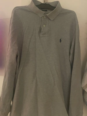 Grå långärmad piké från Ralph Lauren - Klassisk grå långärmad pikétröja från Ralph Lauren. Skjortan har krage, knappar. Säljer min brors tröja som verkar vara i bra skick. 