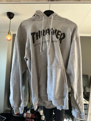 Grå Thrasher hoodie med tryck - Grå hoodie från Thrasher med svart logotyptryck på bröstet. Klassisk känguruficka framtill och justerbar huva med snörning. Tillverkad i mjuk bomullsmix för skön och avslappnad stil.