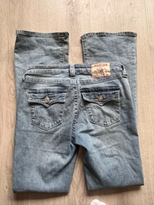 True Religion bootcut jeans  - Säljer ett par ljusblå bootcut jeans från True Religion med klassiska broderade bakfickor och silverfärgade knappar. Jeansen har låg midja och är tillverkade i denim med lätt tvättad look. Perfekta för en avslappnad och trendig stil.