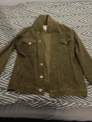 Olivgrön manchesterjacka  - Säljer en olivgrön manchesterjacka med klassisk krage och knappstängning. Jackan har två bröstfickor med lock och knappar samt två sidofickor. Perfekt för dig som gillar retrovibbar och vill ha en unik look.