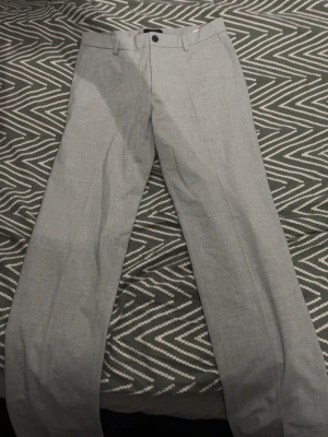 Två par kostymbyxor i grått och beige - Säljer två par stilrena kostymbyxor, ett par i ljusgrått och ett par i beige. Båda har raka ben, pressveck och hällor för bälte. Perfekta för en clean och klassisk look. Materialet känns lätt och passar bra till skjorta eller kavaj.