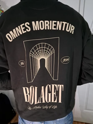 Svart hoodie från Bolaget - Säljer en svart hoodie från Bolaget med trycket 'OMNES MORIENTUR' och grafiskt motiv på ryggen samt 'BR' på bröstet har två fläckar på magen men syns knappt. Hoodien har huva med dragsko och en stor magficka. Perfekt för dig som gillar streetwear och snygga detaljer.