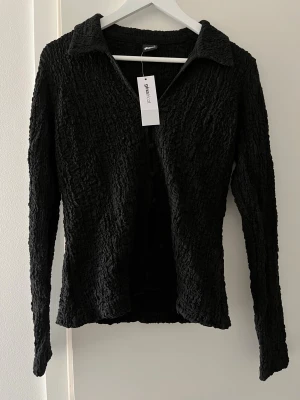 Svart texturerad topp från Gina Tricot - Säljer en svart långärmad topp från Gina Tricot med krage och texturerad, skrynklig yta. Toppen har knappar framtill och en smickrande passform som passar perfekt till olika stilar. Enkel att matcha och supertrendig!