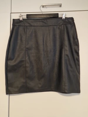 Svart kortkjol i skinnimitation - Snygg svart kortkjol i skinnimitation med enkel och stilren design. Kjolen har raka sömmar framtill och baktill och stängs med dragkedja i sidan. Perfekt att matcha med en cool t-shirt eller stickad tröja.