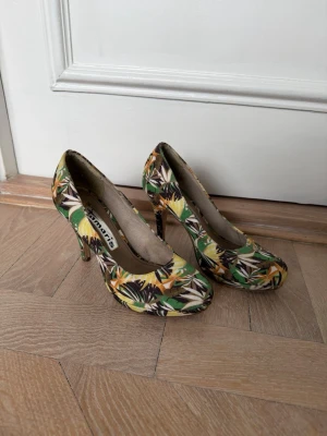 Blommiga pumps från Tamaris - Säljer ett par färgglada pumps från Tamaris med blommigt mönster i grönt, gult, vitt och brunt. Skorna har rundad tå och smal klack, perfekta för dig som vill sticka ut. Materialet är textil med tryckt mönster.