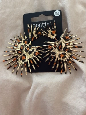 Guldiga Statementörhängen med leopardmönster - Säljer ett par stora statementörhängen från Montini (ur&penn) i metall med leopardmönster i beige, svart och orange. Örhängena har en unik solfjäderform med spretiga detaljer.  