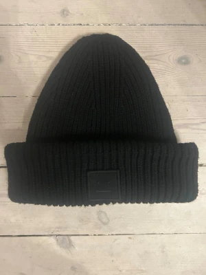 Svart ribbstickad mössa Acne Studios - Svart ribbstickad mössa från Acne Studios med uppvikt kant och den klassiska Acne-loggan framtill. Tillverkad i mjukt och varmt material som håller dig bekväm under kalla dagar. Enkel och stilren design som passar till det mesta. Köpt på plick men kommer inte till användning.