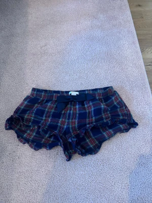 Rutiga pyjamasshorts med volang - Supersöta pyjamasshorts i mörkblått rutigt mönster med röda och gröna detaljer. Shortsen har volangkant och en dekorativ rosett framtill i midjan. Perfekta för mysiga kvällar hemma.