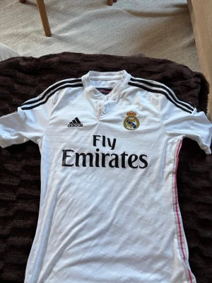 Real Madrid matchtröja Adidas vit - Säljer en vit Real Madrid matchtröja från Adidas med svarta detaljer och klubbmärke på bröstet. Tröjan har Fly Emirates-tryck på framsidan, LFP-märke på ärmen och klassiska Adidas-ränder på axlarna. Perfekt för fotbollsfans!