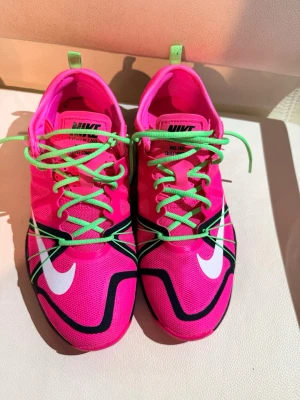 Nike rosa sneakers med gröna snören - Färgstarka sneakers från Nike i neonrosa mesh med vita och svarta detaljer. Skorna har gröna snören och en unik sula med hexagonmönster i svart och rosa. Perfekta för dig som vill ha något som sticker ut och ger energi till din outfit.