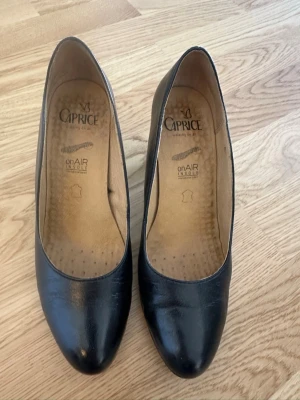 Svarta pumps från Caprice i skinn stl 39 - Stilrena svarta pumps från Caprice med rundad tå och klassisk design. Skorna är tillverkade i äkta skinn och har en bekväm onAIR innersula för extra komfort. Perfekta för dig som vill ha en tidlös och elegant look. 6 cm klack