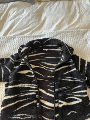 Svartvit teddyjacka med zebramönster - Säljer en svart teddyjacka med vitt zebramönster. Jackan har dragkedja framtill, två stora fickor och en hög krage. Materialet är mjukt och fluffigt, perfekt för kyliga dagar. Snygg och trendig design som sticker ut.