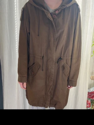Brun parkas med huva och dragsko - Säljer en brun parkas med huva och dragsko i midjan. Fint skick! Från Whyred