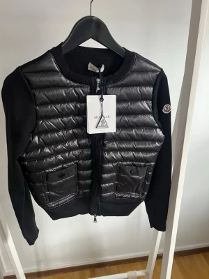 Svart cardiganjacka från Moncler - Snygg svart cardiganjacka från Moncler med quiltad framsida i glansigt material och två fickor med knappar. Ärmar och baksida i mjuk stickad bomull. Rund halsringning och dragkedja framtill. Perfekt för lager-på-lager-stil.