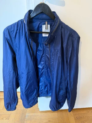 Blå vindjacka från J.Lindeberg - Snygg blå vindjacka från J.Lindeberg med dragkedja framtill och hög krage. Tillverkad i lätt och tunt material som passar perfekt för blåsiga dagar. Jackan har elastiska muddar vid ärmslut och nederkant för extra komfort.