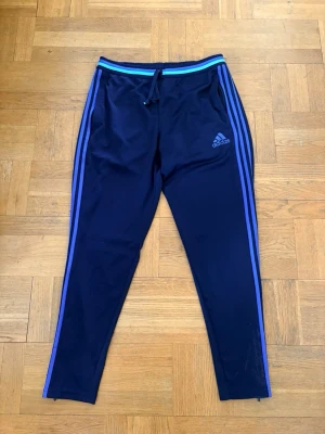 Blå träningsbyxor från Adidas - Säljer ett par mörkblå träningsbyxor från Adidas med blå ränder längs benen och dragsko i midjan. Byxorna har Adidas-logga på låret och är tillverkade i ett lätt, ventilerande material (climacool). Perfekta för träning eller chill. Nypris runt 500kr mitt pris 300kr
