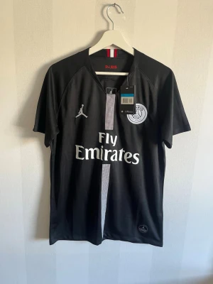 Svart PSG x Jordan t-shirt - Snygg svart PSG t-shirt i samarbete med Jordan, med vit logga och texten 'Fly Emirates' på bröstet. T-shirten har korta ärmar, rund hals och en vertikal rand i mitten. Tillverkad i lätt och skönt polyestermaterial, perfekt för fotboll eller streetwear.